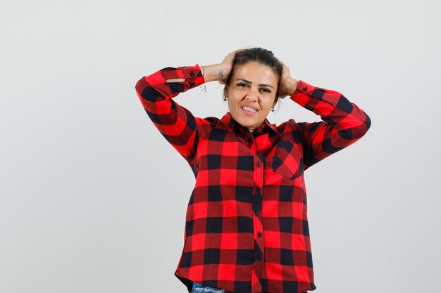 Señorita en camisa a cuadros tomados de la mano en la cabeza y mirando feliz