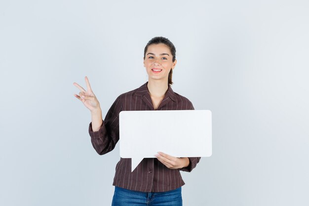 Señorita en camisa, jeans mostrando el signo V, manteniendo un cartel de papel y luciendo feliz, vista frontal.