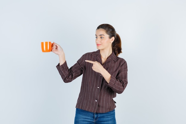 Señorita en camisa, jeans mostrando taza, apuntando hacia un lado y mirando complacido, vista frontal.