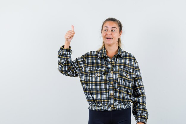 Señorita en camisa, pantalones cortos mostrando el pulgar hacia arriba y mirando feliz, vista frontal.