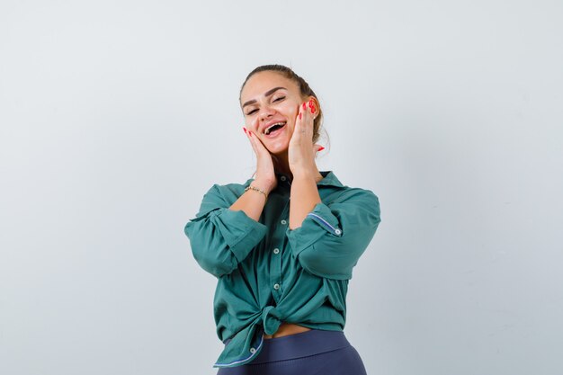 Señorita en camisa, pantalones con palmas en las mejillas y mirando alegre, vista frontal.