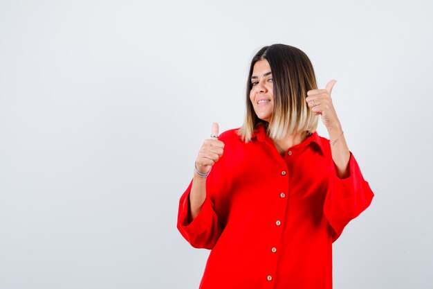 Señorita en camisa roja de gran tamaño mostrando los pulgares hacia arriba y mirando alegre, vista frontal.