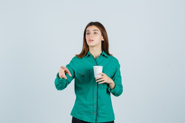 Señorita en camisa sosteniendo una taza de café de plástico, apuntando hacia otro lado y mirando enfocado, vista frontal.