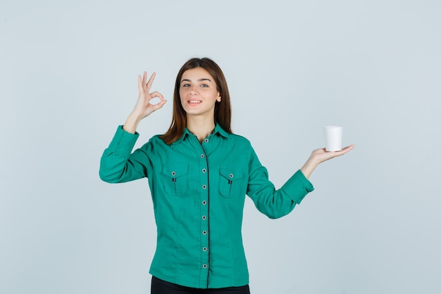 Señorita en camisa sosteniendo una taza de café de plástico mientras muestra un gesto de ok y luce alegre, vista frontal.