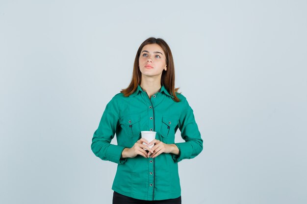 Señorita en camisa sosteniendo una taza de café de plástico y mirando pensativo, vista frontal.