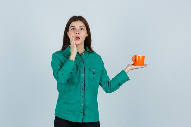 Señorita en camisa sosteniendo una taza de té naranja, manteniendo la mano cerca de la boca y mirando perplejo, vista frontal.