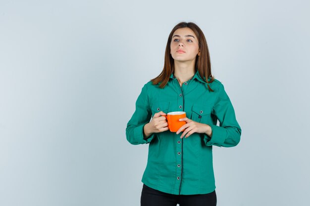 Señorita en camisa sosteniendo una taza de té naranja y mirando confiada, vista frontal.