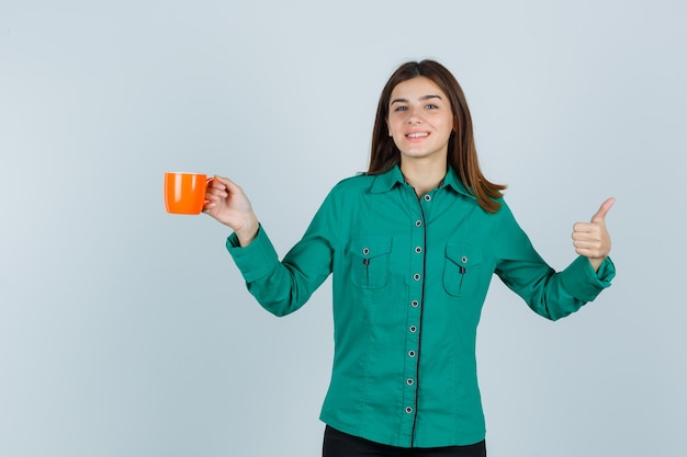 Señorita en camisa sosteniendo una taza de té naranja, mostrando el pulgar hacia arriba y mirando alegre, vista frontal.