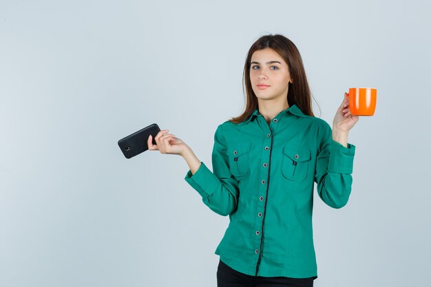 Señorita en camisa sosteniendo una taza de té naranja y un teléfono móvil y mirando confiada, vista frontal.