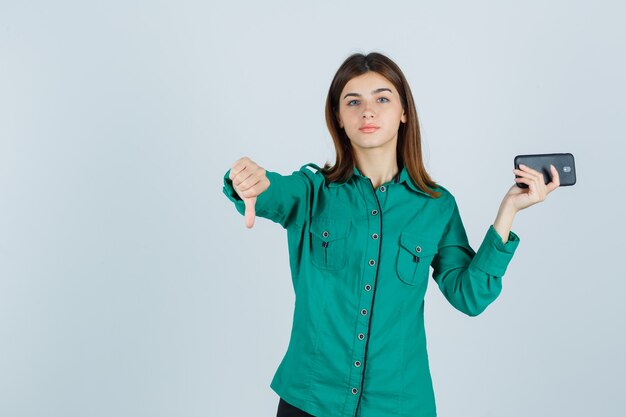 Señorita en camisa verde sosteniendo el teléfono móvil, mostrando el pulgar hacia abajo y mirando disgustado, vista frontal.