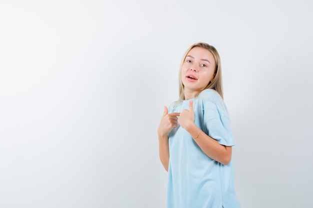 Señorita en camiseta apuntando a sí misma y mirando orgullosa, vista frontal.