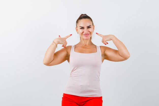 Señorita en camiseta beige apuntando a sí misma y mirando alegre, vista frontal.