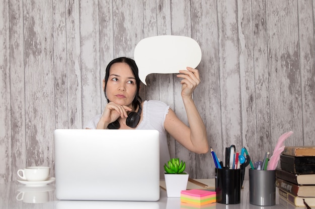 Señorita en camiseta blanca auriculares negros pensando sosteniendo un cartel blanco usando una computadora portátil gris en la mesa taza de café planta bolígrafos libros en gris