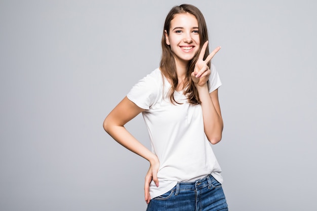 Señorita en camiseta blanca y jeans azul muestra el signo de la victoria frente a fondo blanco de estudio