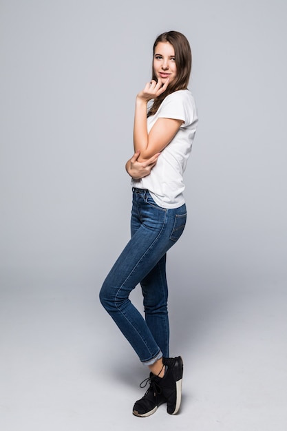 Señorita en camiseta blanca y jeans da un beso al aire delante de un fondo blanco de estudio