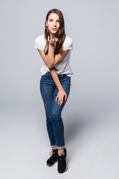 Señorita en camiseta blanca y jeans da un beso al aire delante de un fondo blanco de estudio