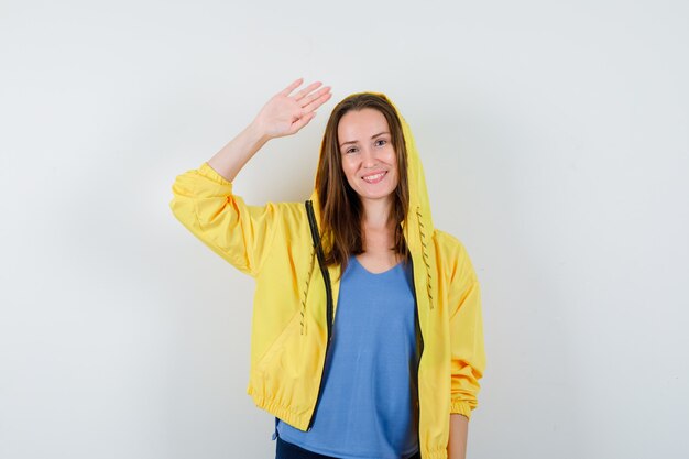 Señorita en camiseta, chaqueta agitando la mano para saludar y mirar feliz, vista frontal.