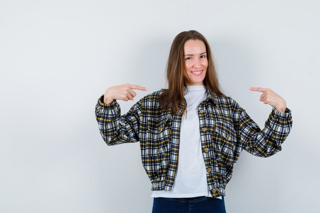 Señorita en camiseta, chaqueta apuntando a sí misma y luciendo orgullosa, vista frontal.