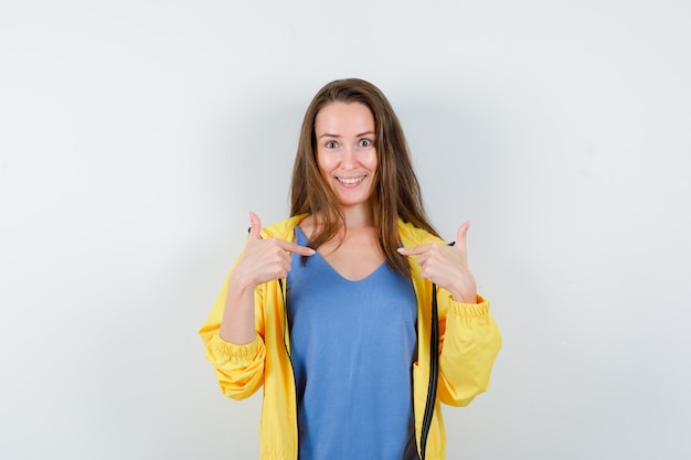 Señorita en camiseta, chaqueta apuntando a sí misma y mirando alegre, vista frontal.