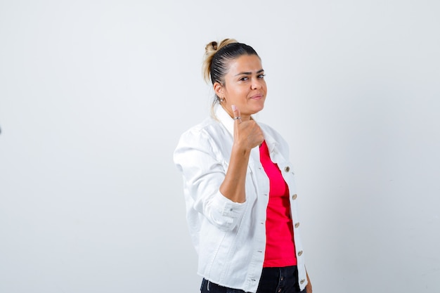 Señorita en camiseta, chaqueta blanca mostrando el pulgar hacia arriba y mirando complacido, vista frontal.