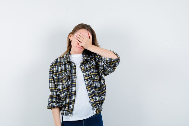 Señorita en camiseta, chaqueta, jeans cubriendo los ojos con la mano y mirando olvidadizo, vista frontal.