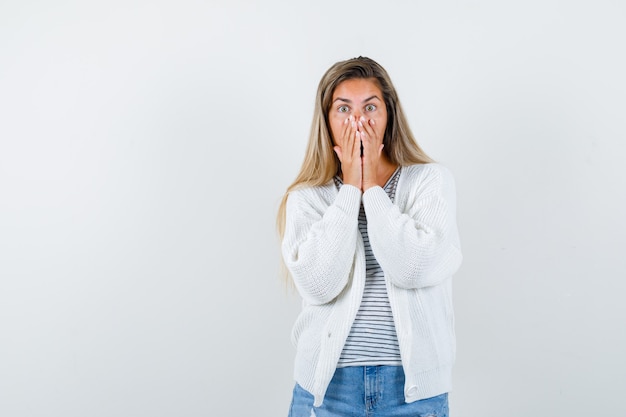 Señorita en camiseta, chaqueta, jeans manteniendo las manos en la boca y mirando sorprendido, vista frontal.