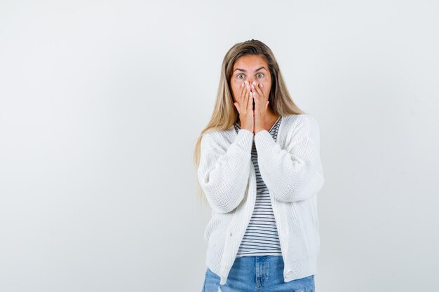 Señorita en camiseta, chaqueta, jeans manteniendo las manos en la boca y mirando sorprendido, vista frontal.