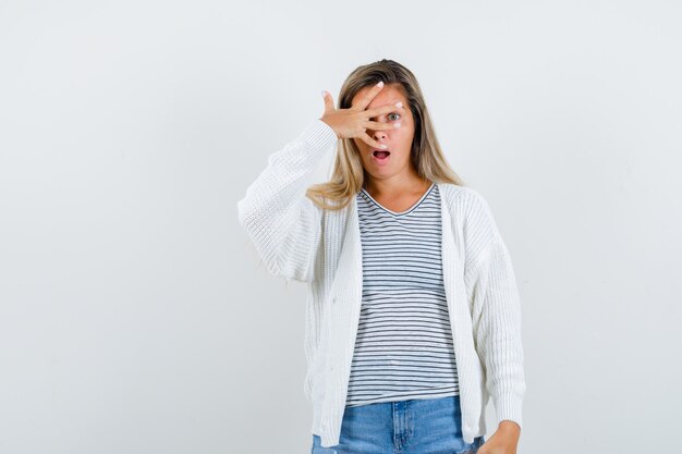 Señorita en camiseta, chaqueta, jeans mirando a través de los dedos y mirando asombrado, vista frontal.