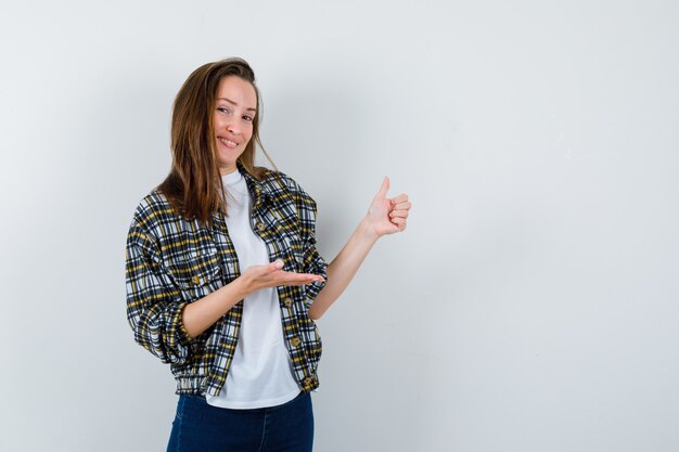 Señorita en camiseta, chaqueta, jeans mostrando el pulgar hacia arriba mientras da la bienvenida a algo y se ve atractiva, vista frontal.