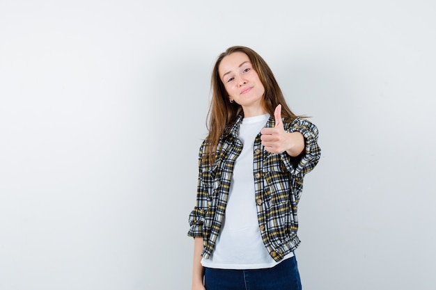 Señorita en camiseta, chaqueta, jeans mostrando el pulgar hacia arriba y mirando confiado, vista frontal.