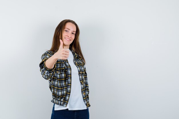 Señorita en camiseta, chaqueta, jeans mostrando el pulgar hacia arriba y mirando confiado, vista frontal.