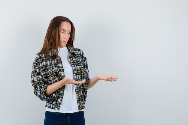 Señorita en camiseta, chaqueta, jeans pretendiendo mostrar algo y mirando perplejo, vista frontal.