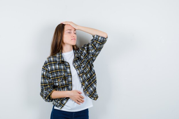 Señorita en camiseta, chaqueta, jeans sosteniendo la mano en la cabeza y mirando olvidadizo, vista frontal.