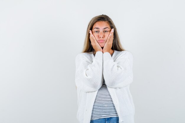 Señorita en camiseta, chaqueta manteniendo las manos en las mejillas y con aspecto cansado, vista frontal.