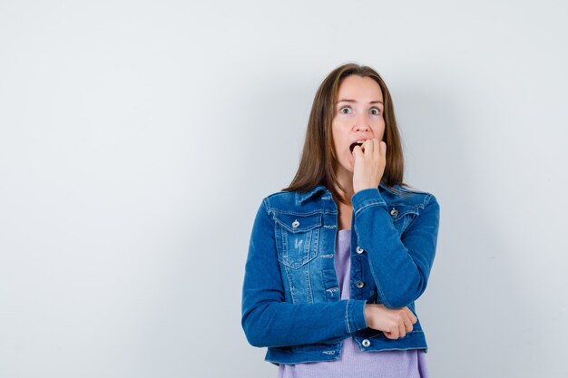 Señorita en camiseta, chaqueta mordiéndose las uñas emocionalmente y mirando ansiosa, vista frontal.