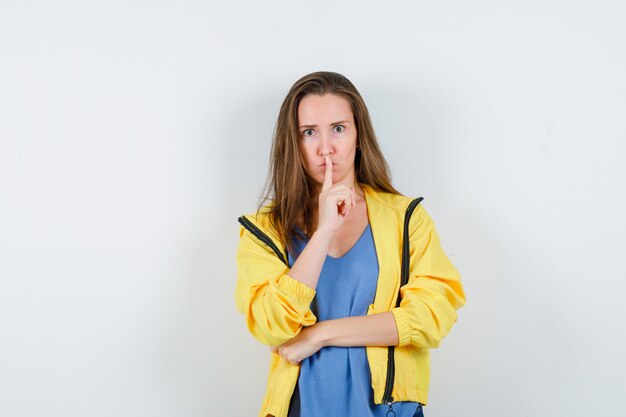 Señorita en camiseta, chaqueta mostrando gesto de silencio y mirando serio, vista frontal.