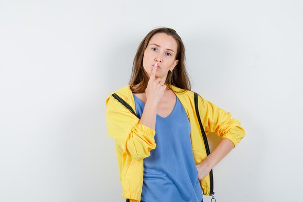 Señorita en camiseta, chaqueta sosteniendo el dedo en los labios y mirando pensativo, vista frontal.