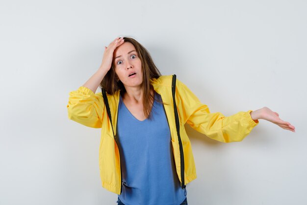 Señorita en camiseta extendiendo la palma a un lado, sosteniendo la mano en la cabeza y mirando desconcertado