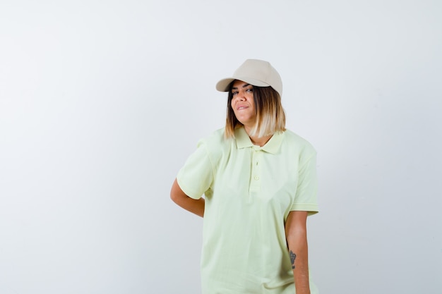 Señorita en camiseta, gorra sosteniendo la mano detrás de la espalda mientras posa y se ve bonita, vista frontal.