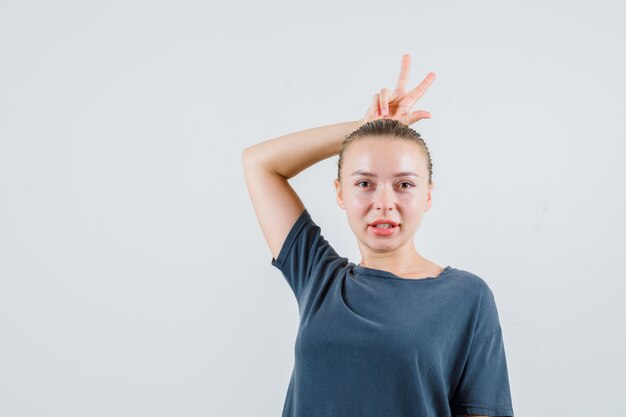 Señorita en camiseta gris que muestra el signo v detrás de la cabeza como cuernos y se ve gracioso