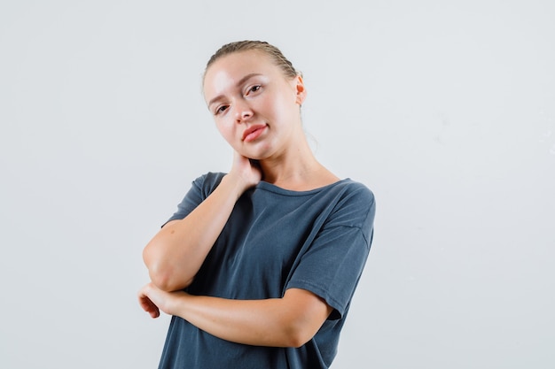 Señorita en camiseta gris sosteniendo la mano en el cuello y mirando linda