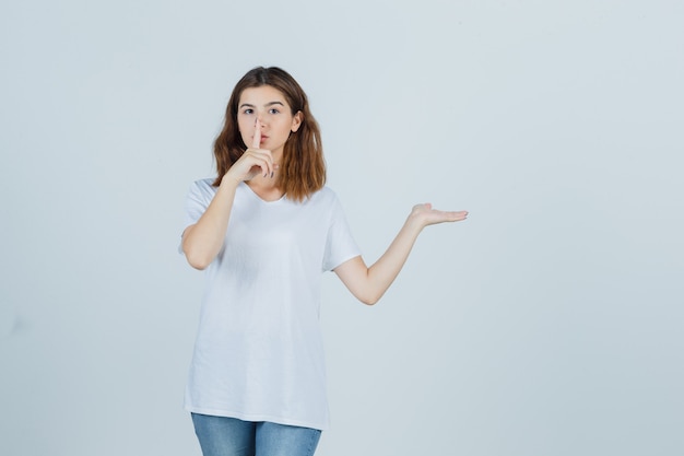 Señorita en camiseta, jeans mostrando gesto de silencio mientras muestra algo y mira seria, vista frontal.