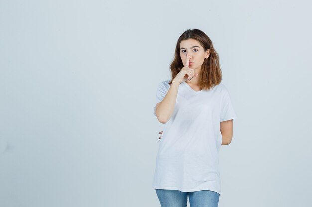 Señorita en camiseta, jeans mostrando gesto de silencio y mirando serio, vista frontal.