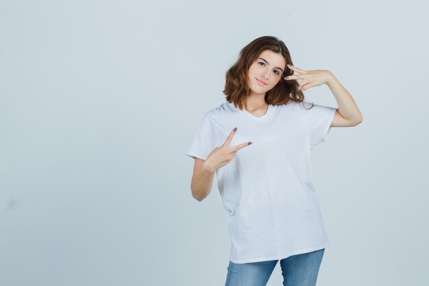 Señorita en camiseta, jeans mostrando el signo de la victoria y mirando alegre, vista frontal.