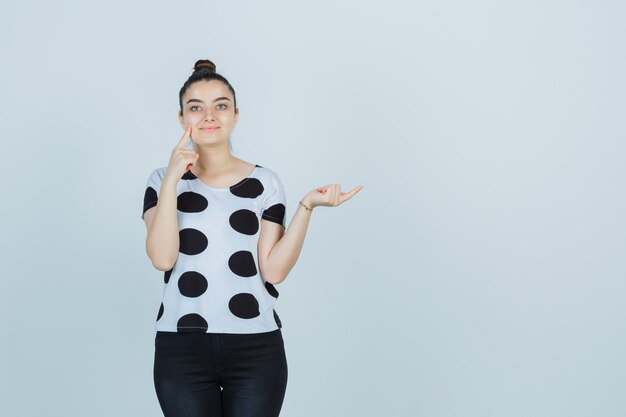 Señorita en camiseta, jeans presionando el dedo en la mejilla mientras apunta hacia el lado derecho y mira confiada, vista frontal.