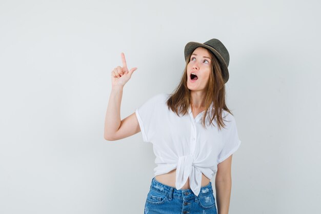 Señorita en camiseta, jeans, sombrero apuntando hacia arriba y mirando asombrado, vista frontal.