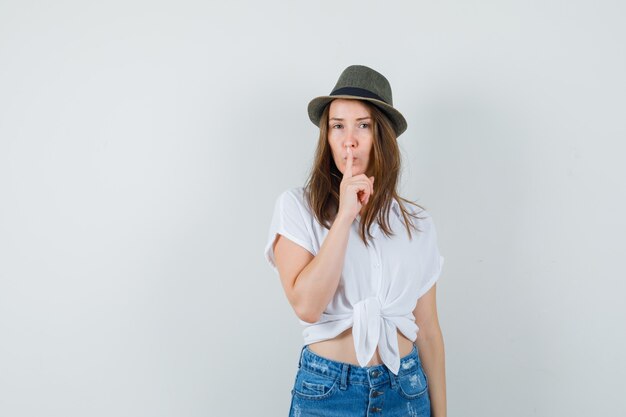 Señorita en camiseta, jeans, sombrero mostrando gesto de silencio y mirando con cuidado, vista frontal.