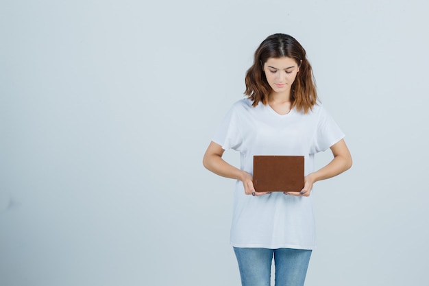 Señorita en camiseta, jeans sosteniendo libro mientras mira y mira pensativa, vista frontal.