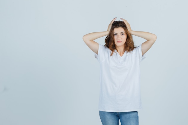 Señorita en camiseta, jeans tomados de la mano en la cabeza y con aspecto cansado, vista frontal.