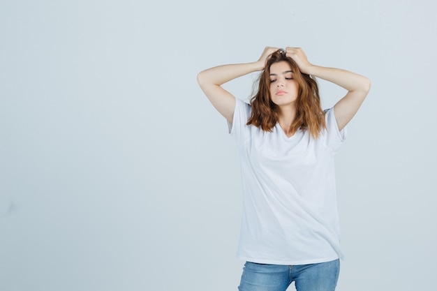 Señorita en camiseta, jeans tomados de la mano en la cabeza y mirando triste, vista frontal.